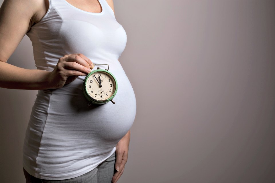 Close up of a pregnant woman's belly, woman standing next to a grey background and holding a clock representing a proximity of a due date