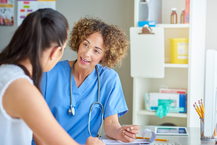 A female doctor sits at her desk and listens to a female patient . She is wearing blue scrubs and stethoscope and is sitting at her desk . In the background shelving containing sharps box, gloves and medicine can be seen .