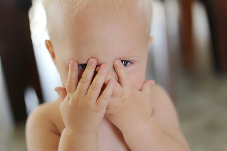 A one year old baby girl is covering her face with her hands and peeking through her fingers.
