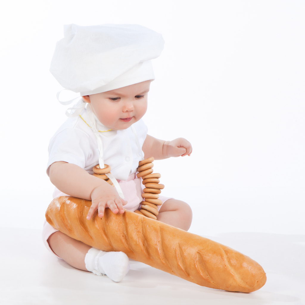 Little baker baby girl with a long loaf and bagels, isolated on white background