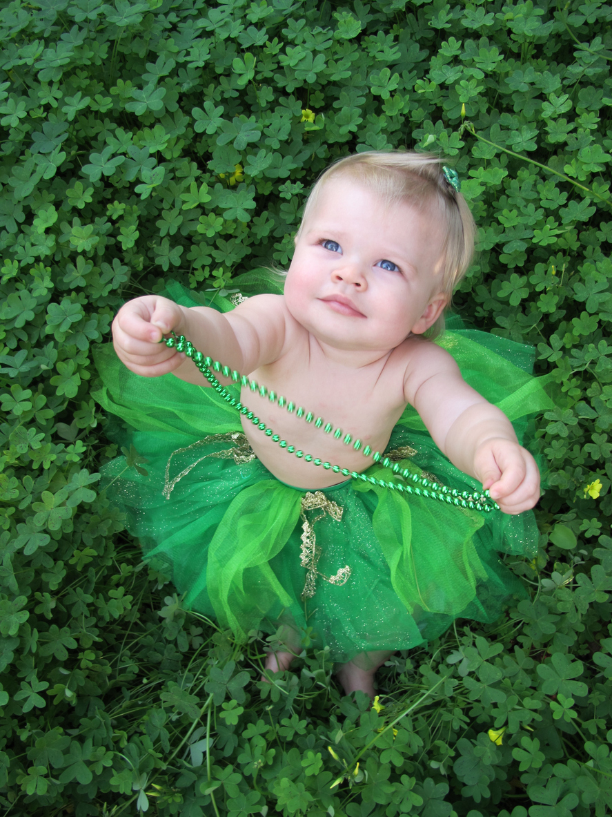 A cute baby girl in a green tutu plays with her green necklace while sitting in a patch of clovers for St. Patrick's Day.