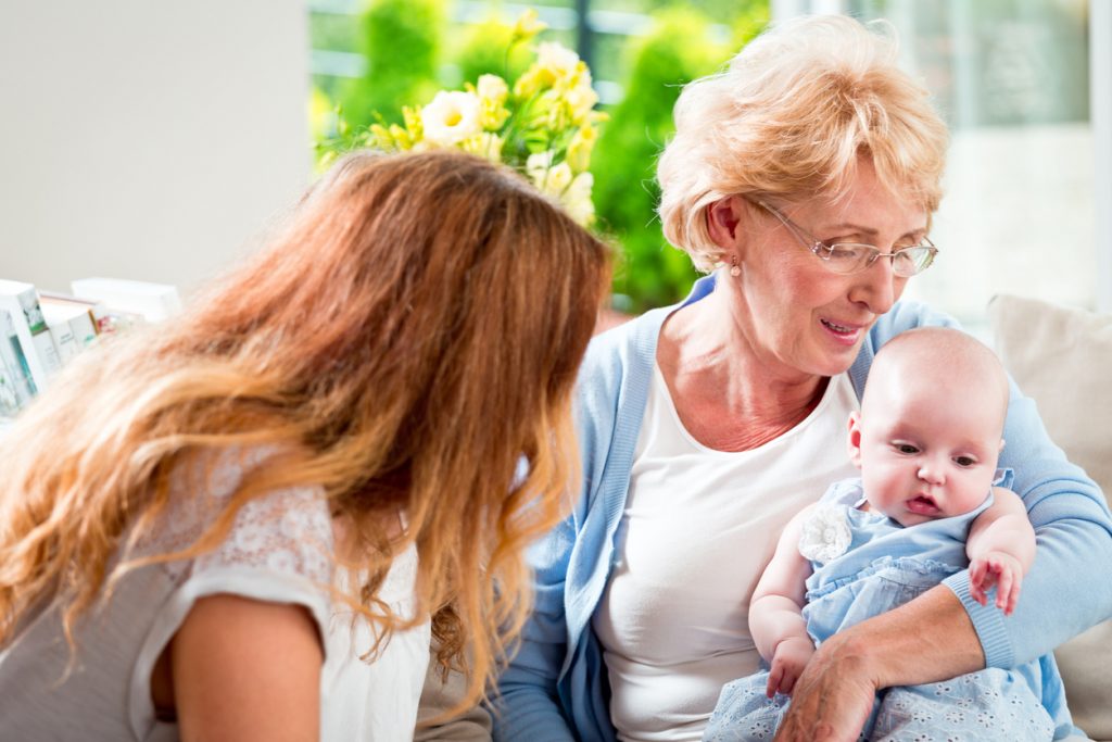 Portrait of happy grandmother embracing her little granddaughter with a woman talking to baby girl.