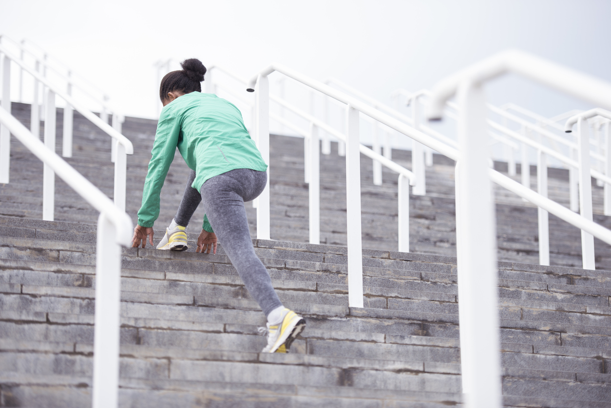 Photo from below of young woman wearing green sport jacket and gray tights, stretching her legs at stadium stairs early in the morning. Fitness, diet, active lifestyle concept.