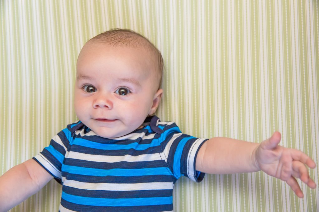 Close-up of a baby boy (two and a half months old) laying in his crib on a green and white striped sheet looking up. The camera is directly above him. The baby is wearing a blue and white striped short sleeve shirt and has a small smile on his face.