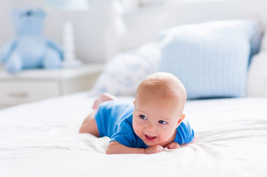 Adorable baby boy in white sunny bedroom. Newborn child relaxing in bed. Nursery for young children. Textile and bedding for kids. Family morning at home. New born kid during tummy time with toy bear.