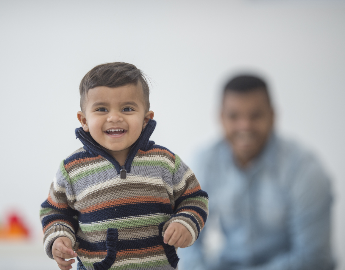 A little boy is playing in his home with his father on father's day. The boy is smiling while looking at the camera.