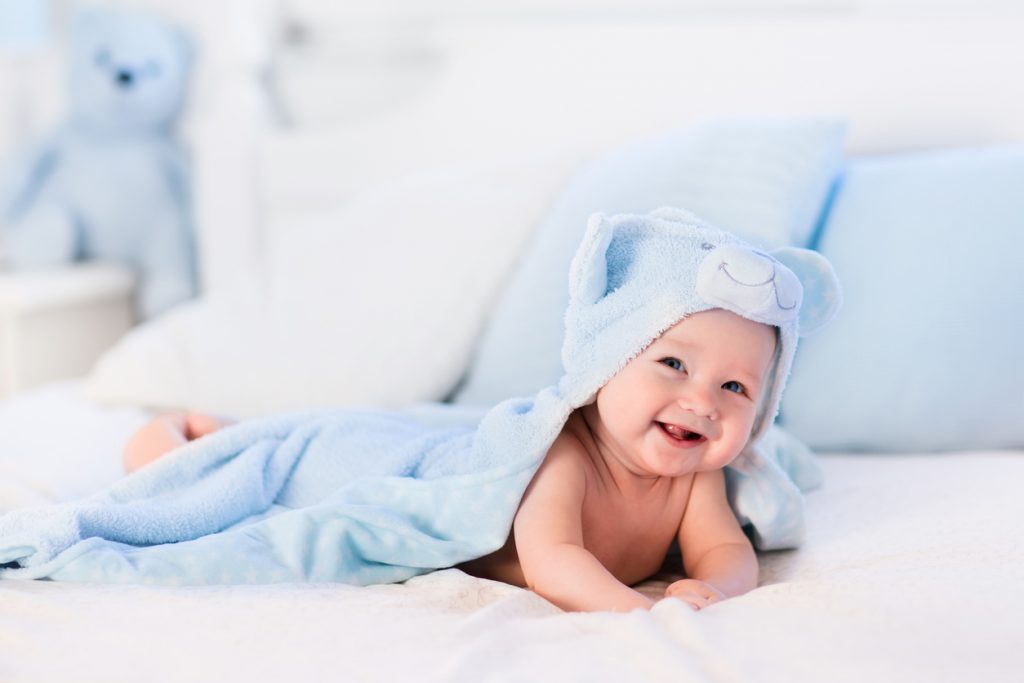 Baby boy wearing diaper and blue towel in white sunny bedroom. Newborn child relaxing in bed after bath or shower. Nursery for children. Textile and bedding for kids. New born kid with toy bear.