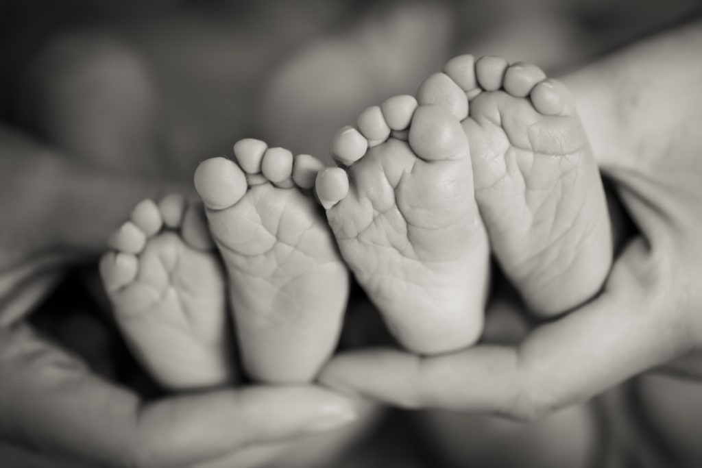 Black and white photo of hands holding four newborn baby twin feet.