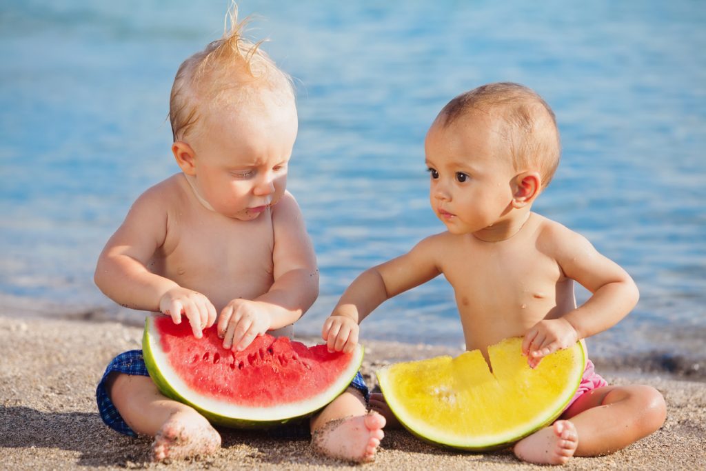 After sea swimming asian and caucasian babies have a fun and eat fresh fruits on sand beach. Healthy kids food, active lifestyle, water activity and children travel with parents on family vacation.