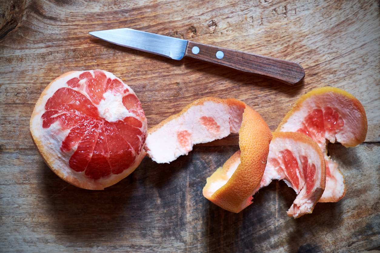 An old-fashioned wooden-handled paring knife sits on a chopping board next to a peeled pink grapefruit.