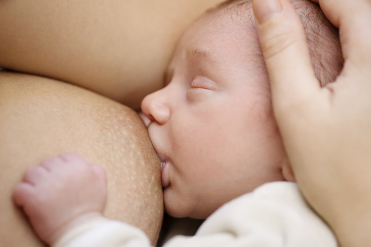 Close-up of a baby breastfeeding. The mother's hand holds his head. Focus on the baby's mouth.