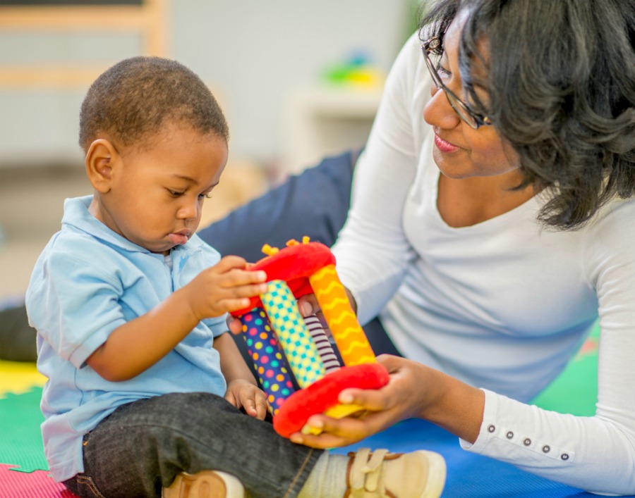 Mother sitting in a preschool classroom with her little boy. They are playing with toys on a mat.