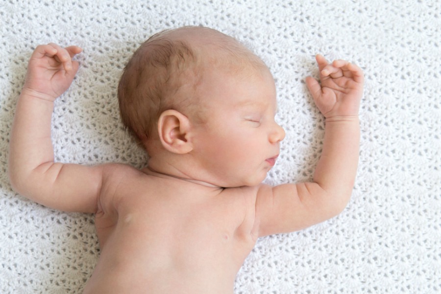 Portrait of young funny newborn babe napping on white knitted blanket with his arms up. Cute caucasian new born child sleeping. Relaxed kid lying on bed with closed eyes. Copyspace. View from above