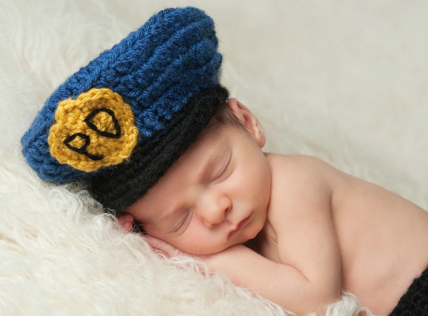 Sleeping 12 day old newborn baby boy wearing a blue and gold crocheted police officer's hat.