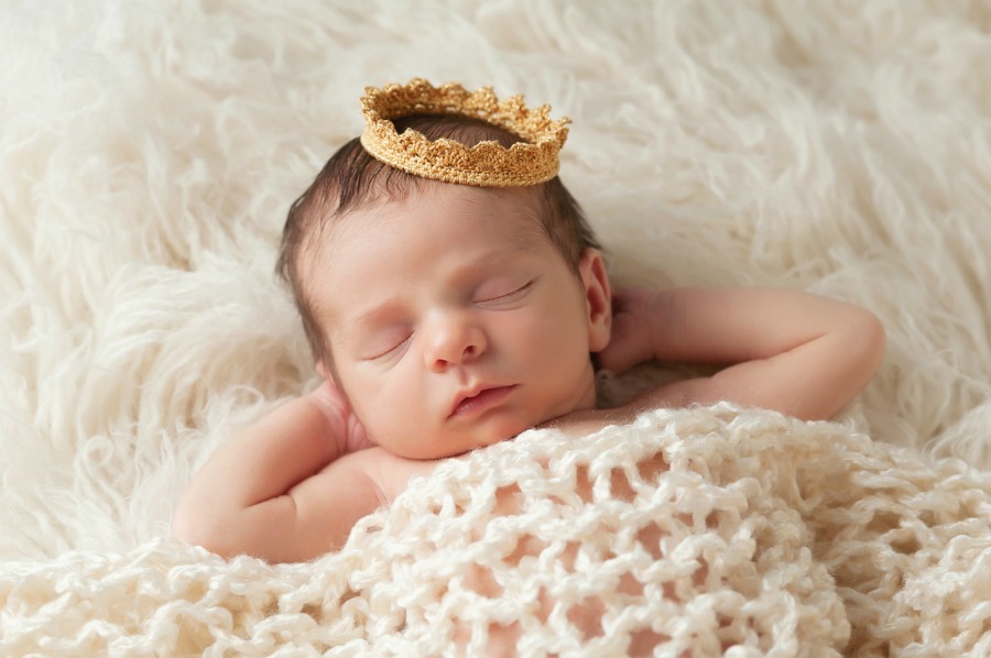 Portrait of a 12 day old newborn baby boy wearing a gold crown. He is sleeping on a beige flokati rug with his hands behind his head.