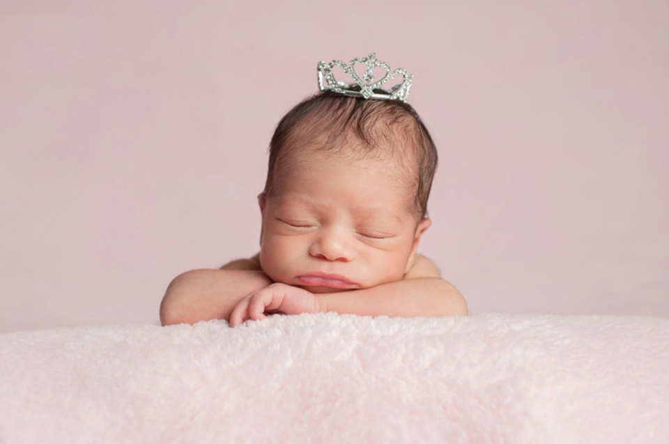 Portrait of two week old sleeping newborn baby girl. She is wearing a rhinestone crown and is posed with her chin on her hands.