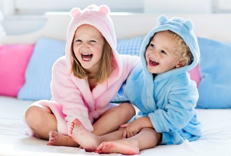 Happy laughing kids, boy and girl in soft bathrobe after bath play on white bed with blue and pink pillows in sunny bedroom. Child in clean and dry towel. Wash, infant hygiene, health and skin care.