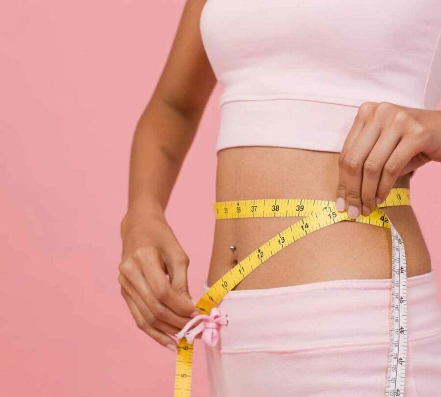 lose weight in 12 weeks - image of a womans waist with peach tights and white top whilst she's measuring her waist using a measuring tape on a pink background